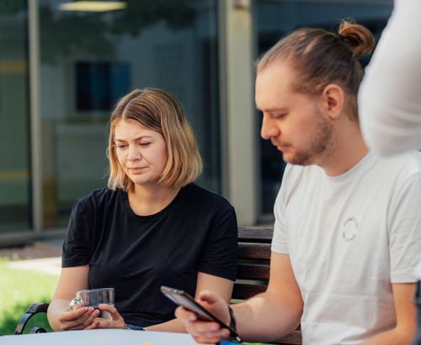 Zwei Personen sitzen draußen am Tisch; eine hält ein Glas, der andere schaut aufs Smartphone.